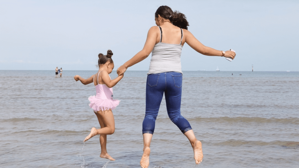 A woman and a young child hold hands and jump together barefoot in the shallow water at the beach