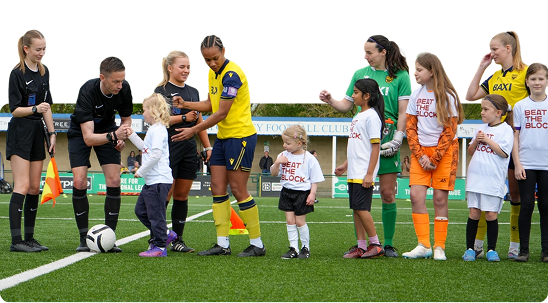 Children and female football players line up on a soccer field before a match, with one child greeting a player.
