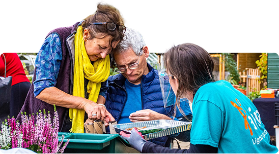 Three people gathered around a gardening tray, examining and planting seedlings together outdoors.
