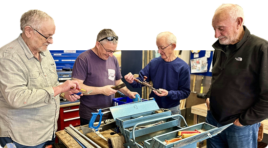 Four older men gathered around a workbench, examining tools and metalwork in a workshop setting.