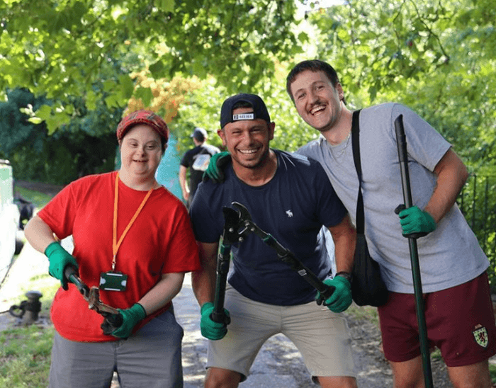 Three smiling volunteers in gardening gloves holding tools on a tree-lined path during an outdoor community task.